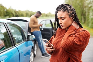 POC woman rubbing neck after car accident with other male driver on the phone in background